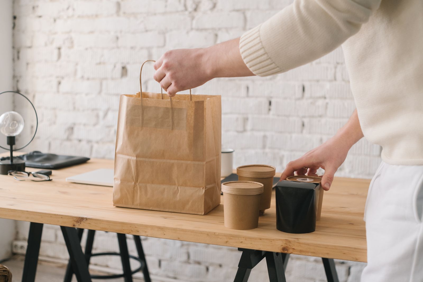 Hands packing takeaway boxes and cups into a paper bag on a wooden counter