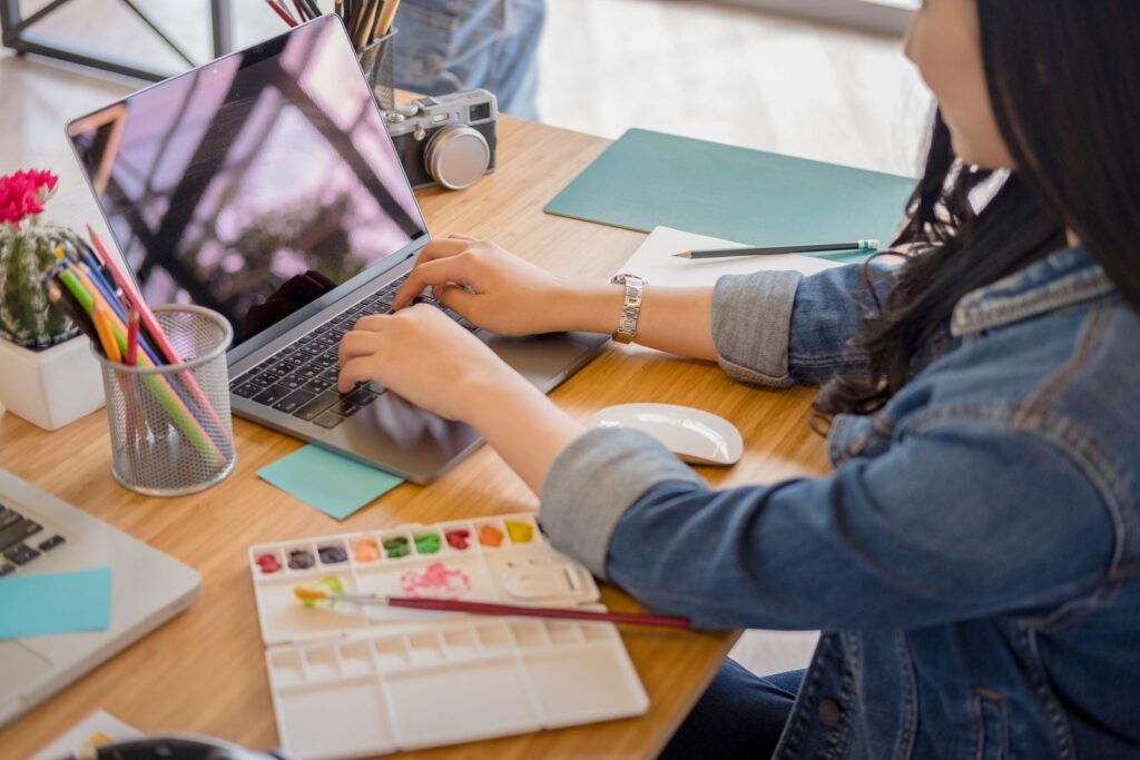 Hands typing on a laptop at a desk with a notebook and pencils