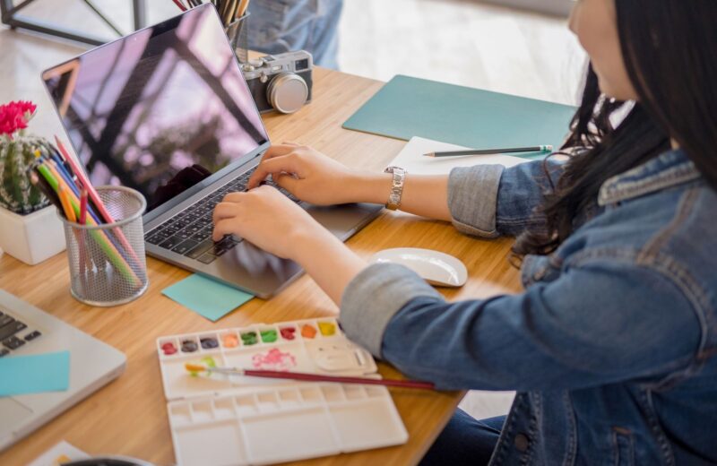 Hands typing on a laptop at a desk with a notebook and pencils