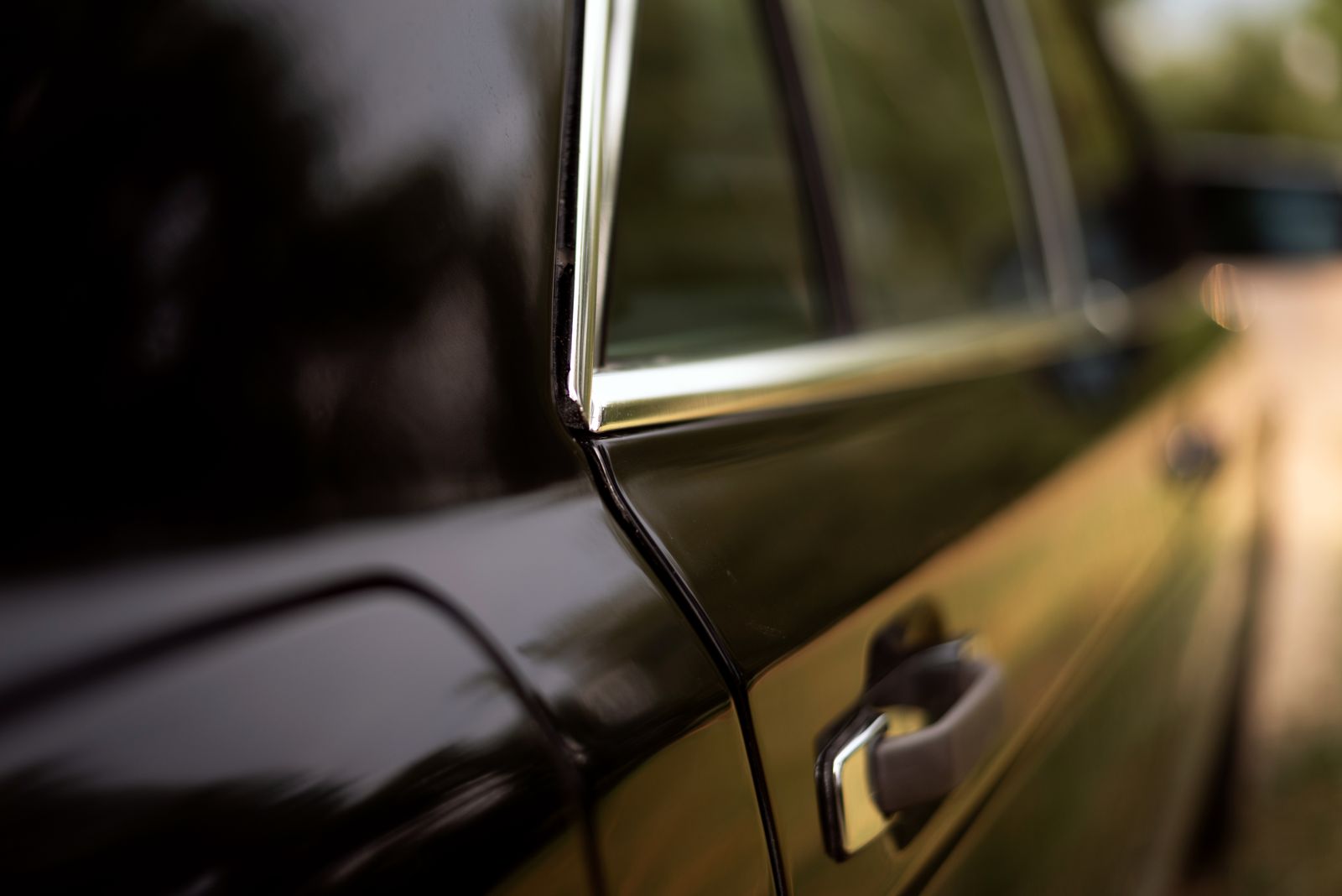 The black door of a classic car with a chrome handle and window trim, light running along the paint
