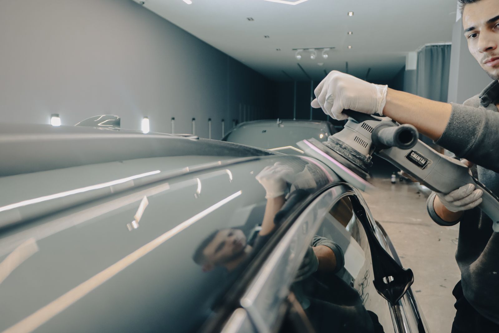 A detailer in gloves polishing the roof of a grey car with a machine polisher in a white studio