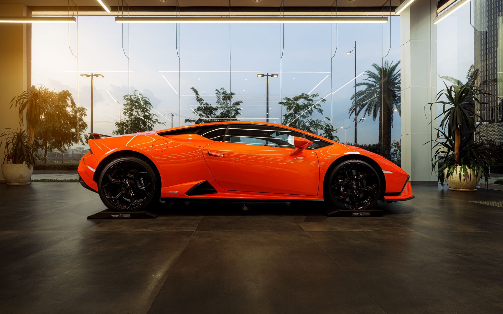 An orange sports coupe seen side-on in a glass showroom, palm trees outside the windows