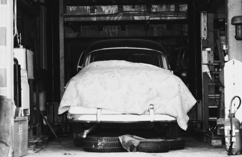 Black and white photo of a classic car under a fitted cover in a garage