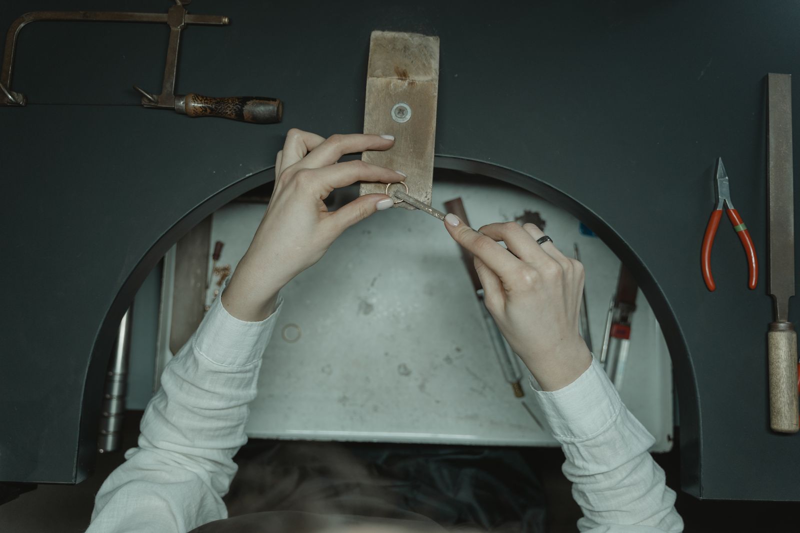 Hands at a jeweller's bench seen from above, filing a ring on the bench peg among pliers and files
