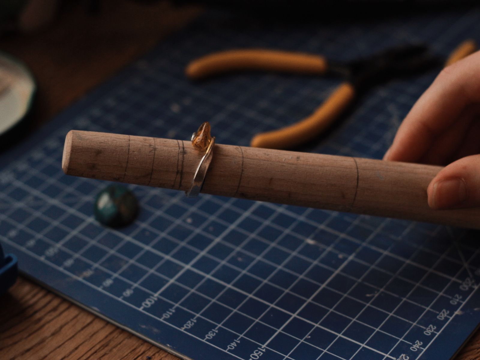 A hand holding a ring on a wooden ring mandrel above a cutting mat, with pliers behind