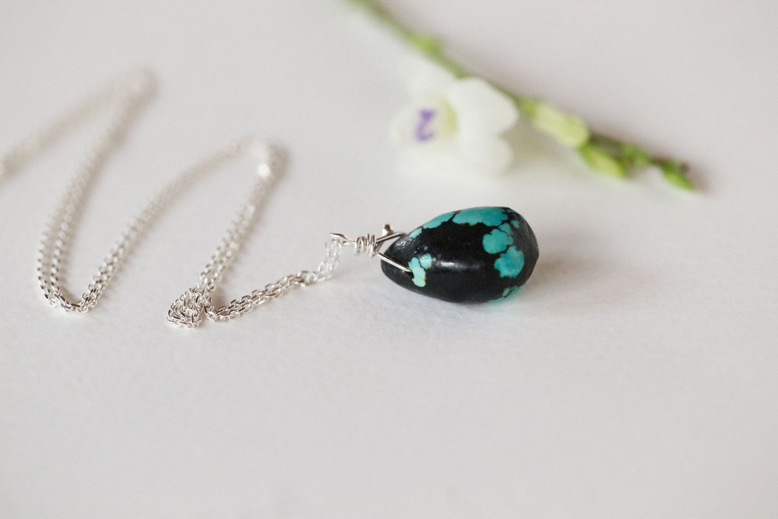 A silver chain with a green glass pendant on a pale table beside a white flower