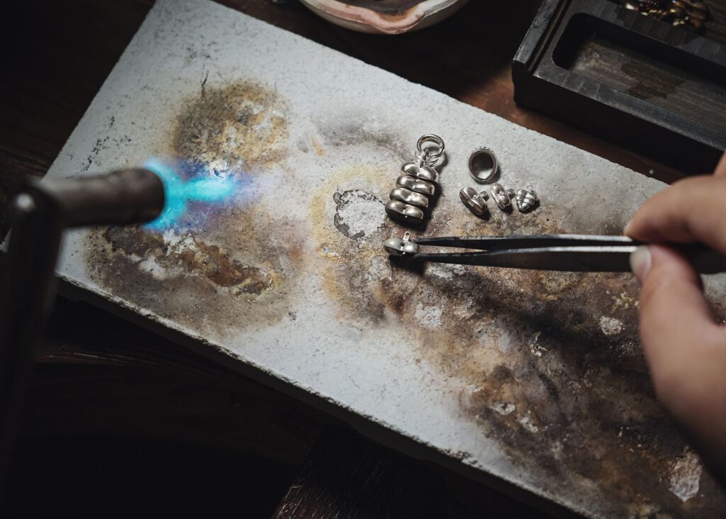 A torch flame and tweezers on a scorched soldering block with small silver parts