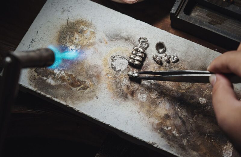 A torch flame and tweezers on a scorched soldering block with small silver parts
