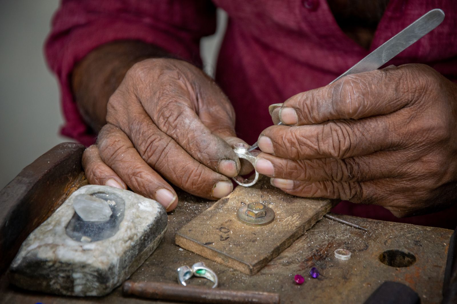 Weathered hands setting a stone in a silver ring at an old wooden bench, with loose gems beside a soldering block