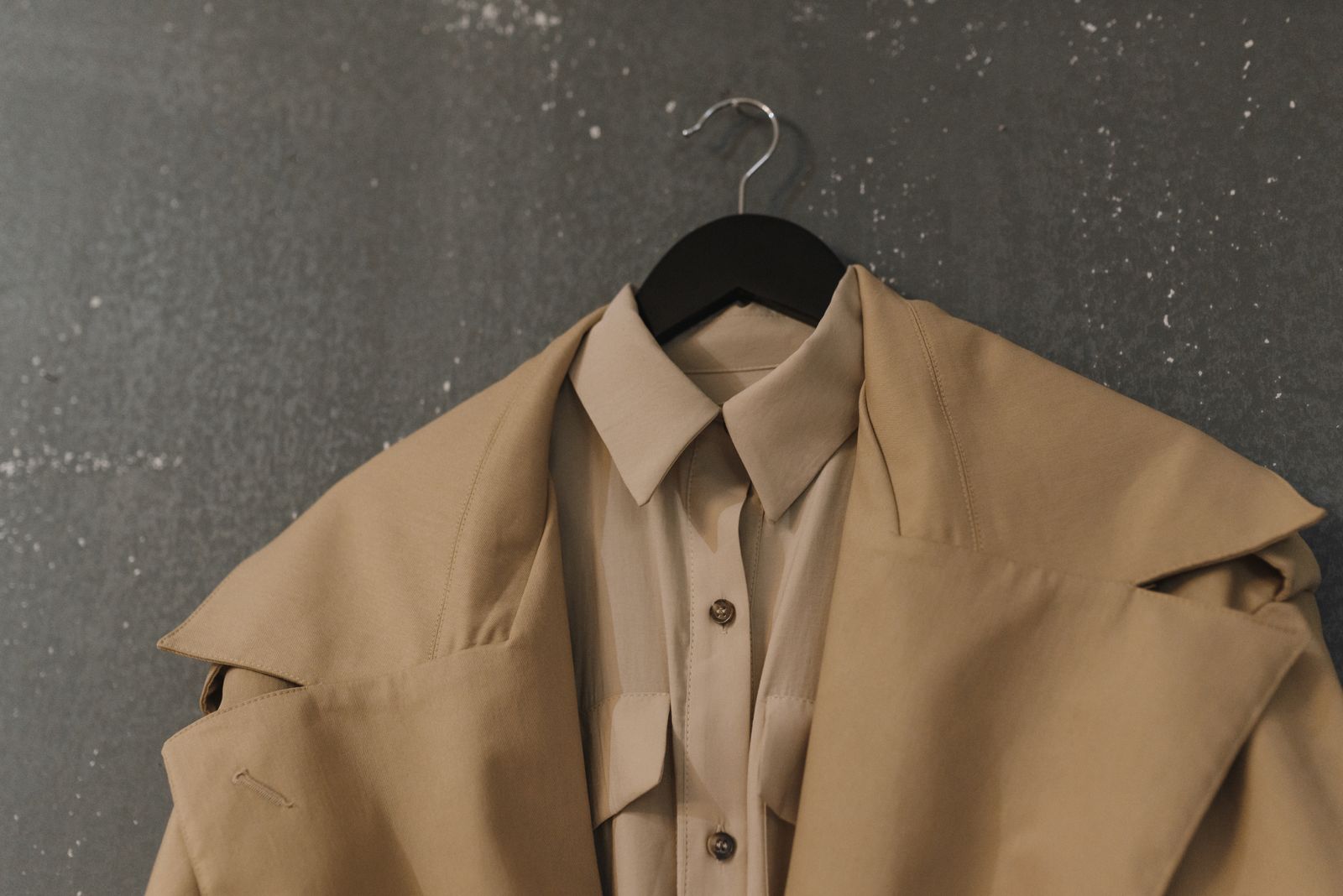 A camel coat over a shirt on a hanger against a grey wall