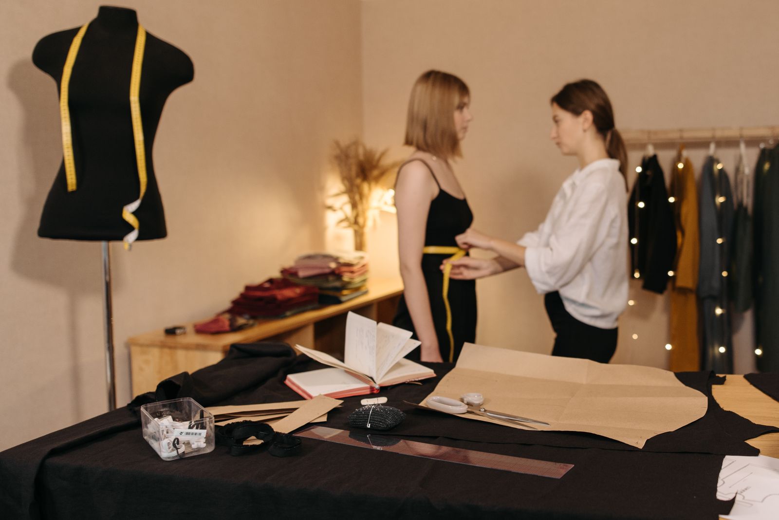 A tape measure round a customer's waist in a fitting room, with a dress form and pattern paper on the table