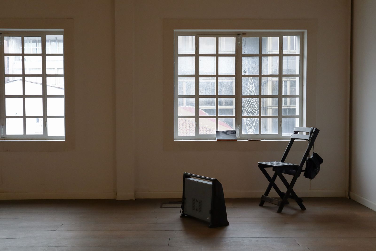 An empty room with tall white-framed windows, a dark folding chair and a small heater on a wooden floor