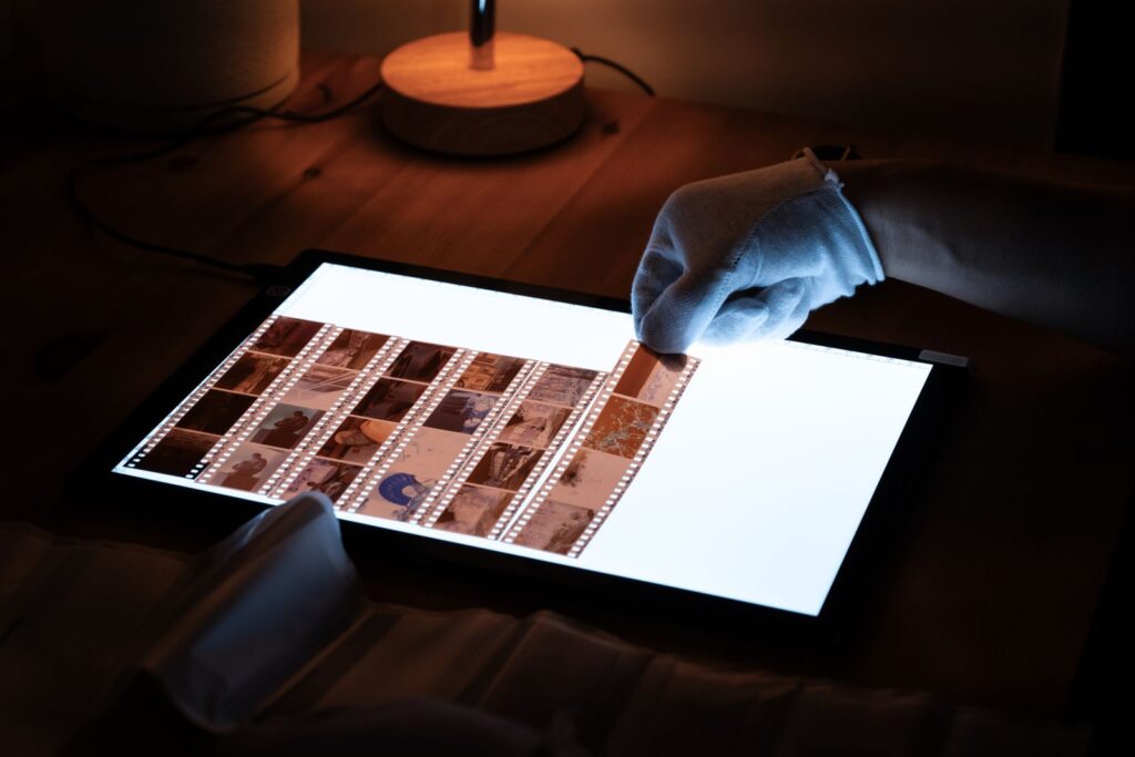 A gloved hand turning strips of film negatives on a lit light table
