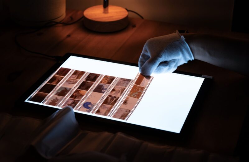 A gloved hand turning strips of film negatives on a lit light table