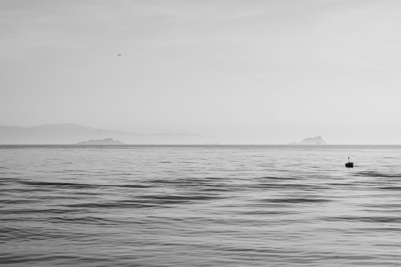 A grey sea under a pale sky with distant islands and a small buoy, in black and white