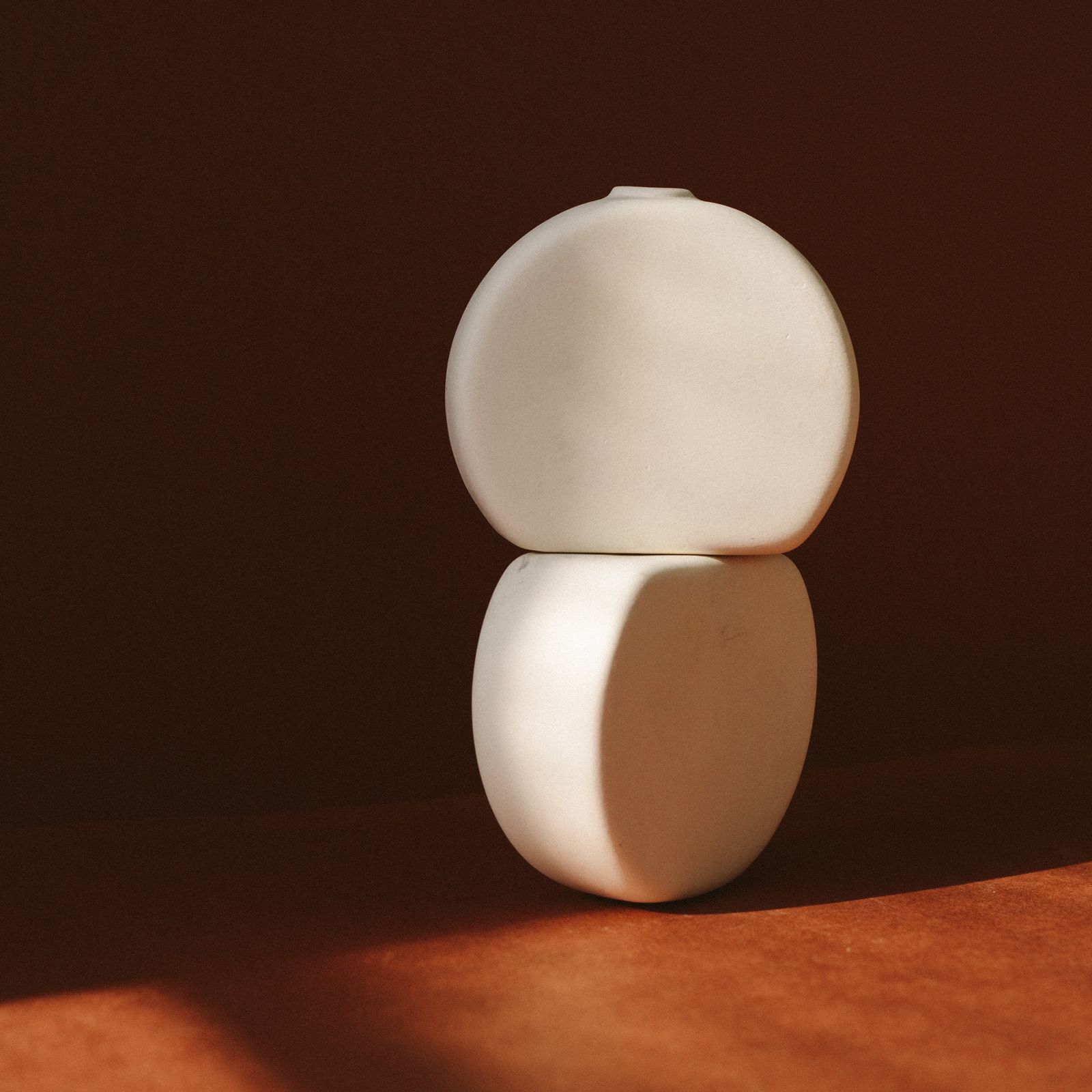 A round white ceramic vase in low warm sunlight against a brown wall