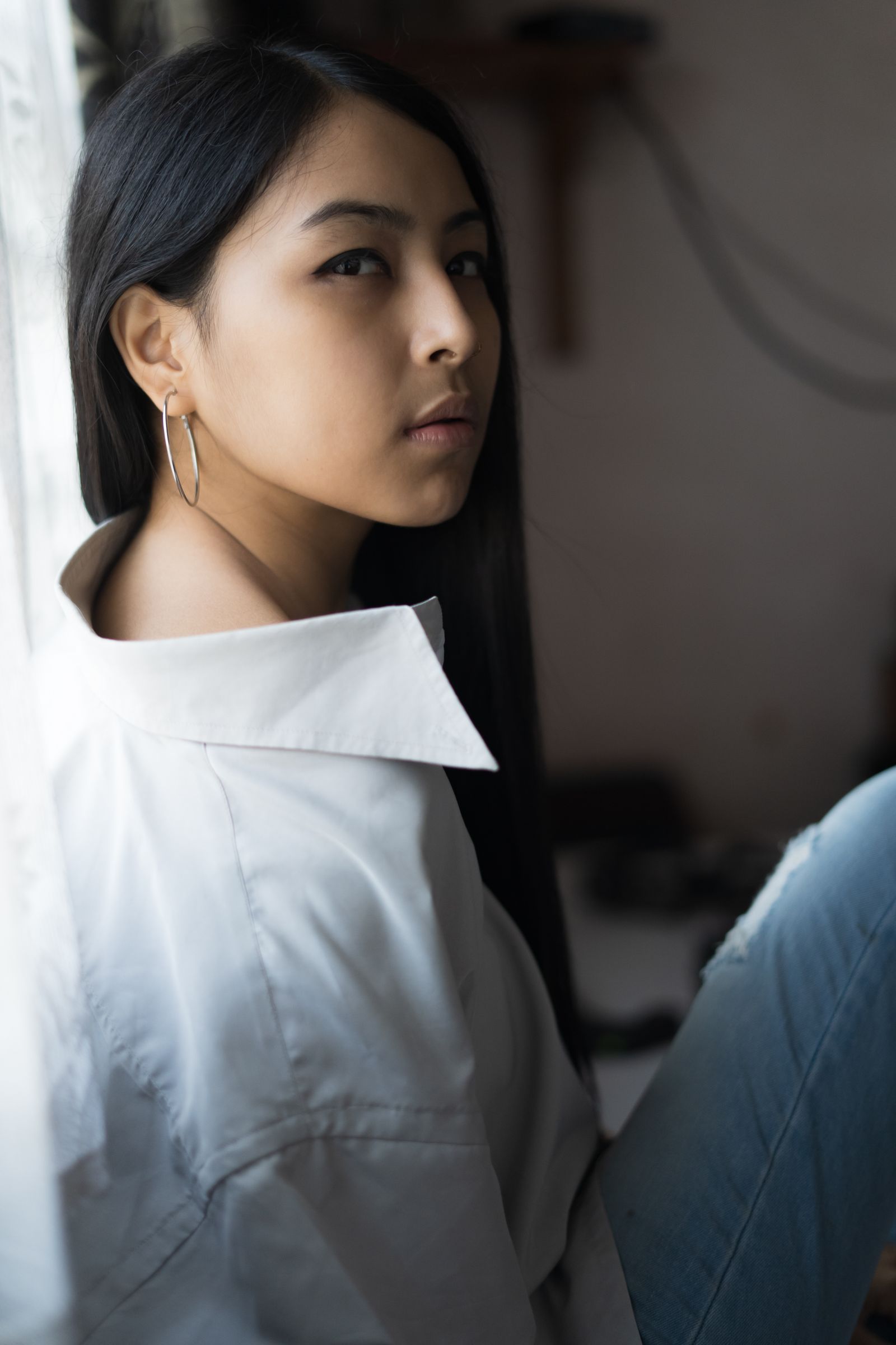 A woman in a white shirt sitting by a window, her face turned away and partly out of frame