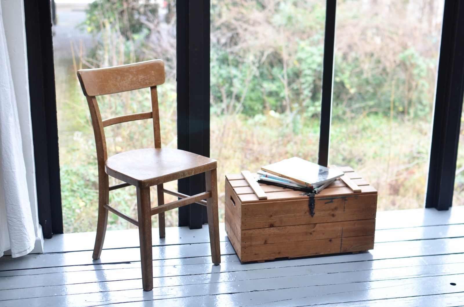A wooden chair and a crate with books in front of a black-framed glass wall and a garden