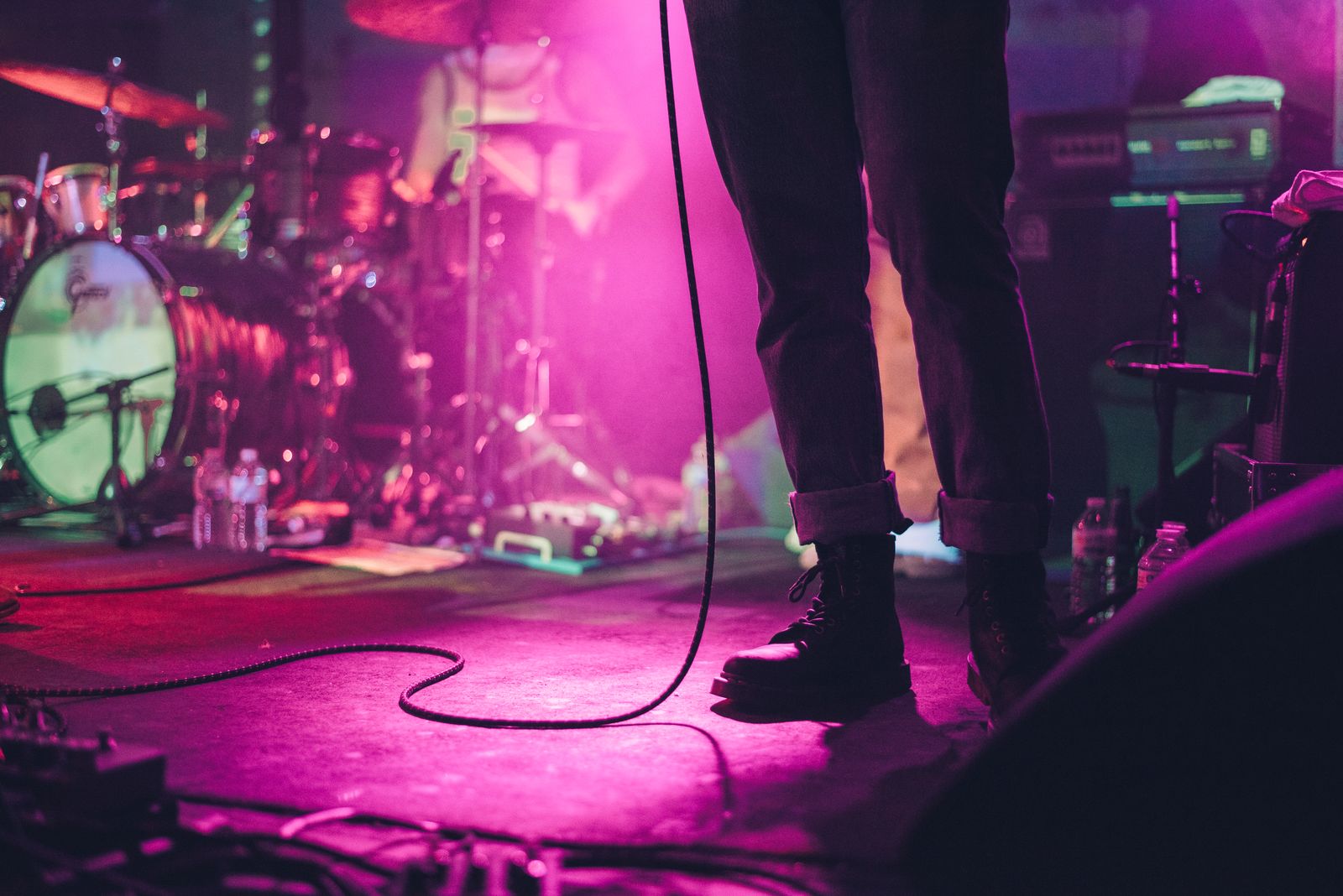 A band on a small stage under pink and violet light, the singer's boots and a microphone cable in front of the drum kit