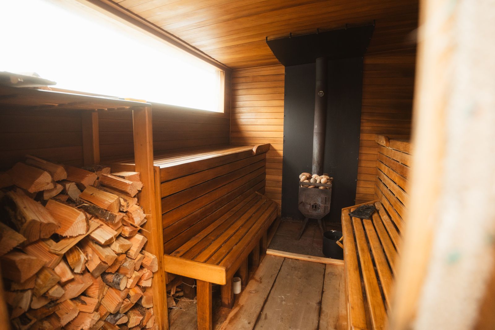 A wood-fired sauna with a black stove, slatted benches and a stack of split logs