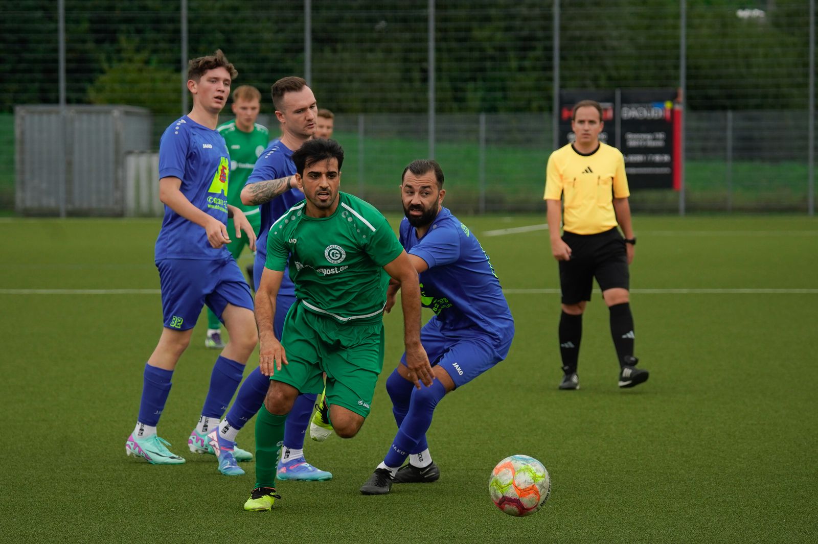 Footballers in green and blue chasing the ball on a grass pitch, the referee watching