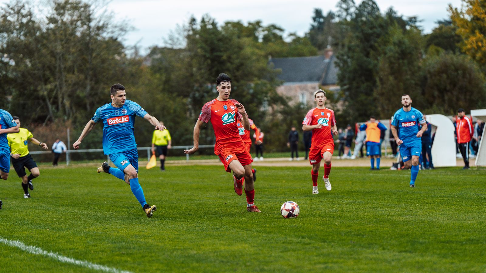 A football match on a wide grass pitch, players in red and in blue running for the ball