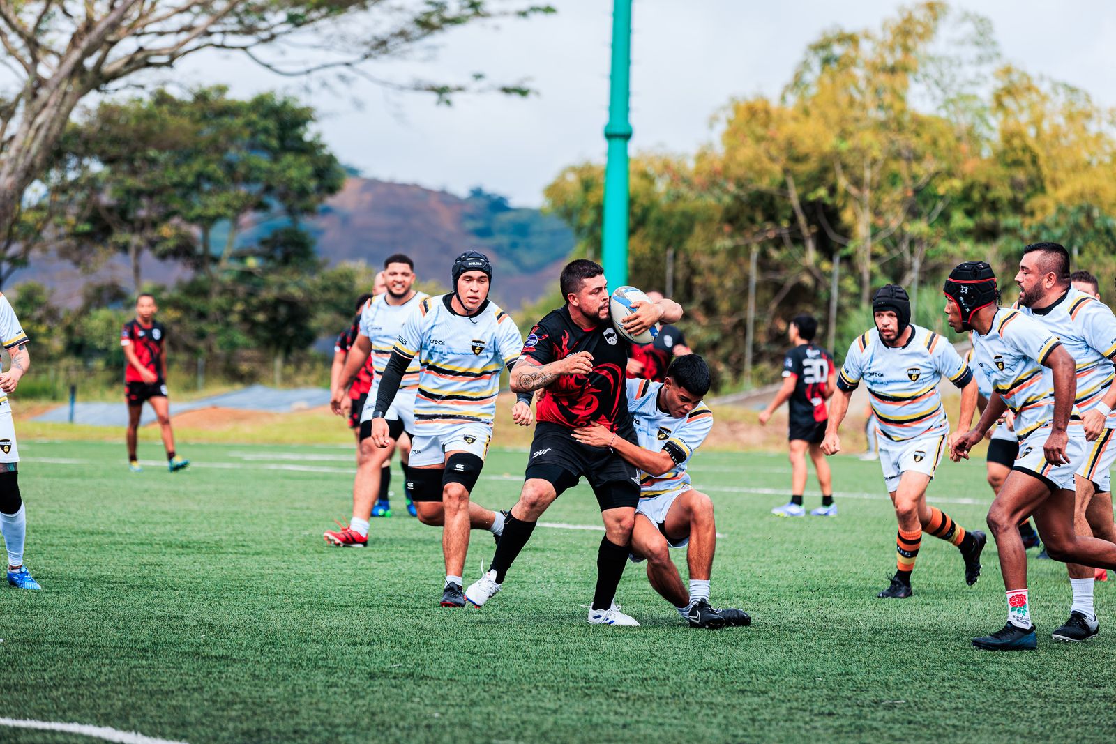 A rugby match seen from the touchline, players spread across a sunny pitch