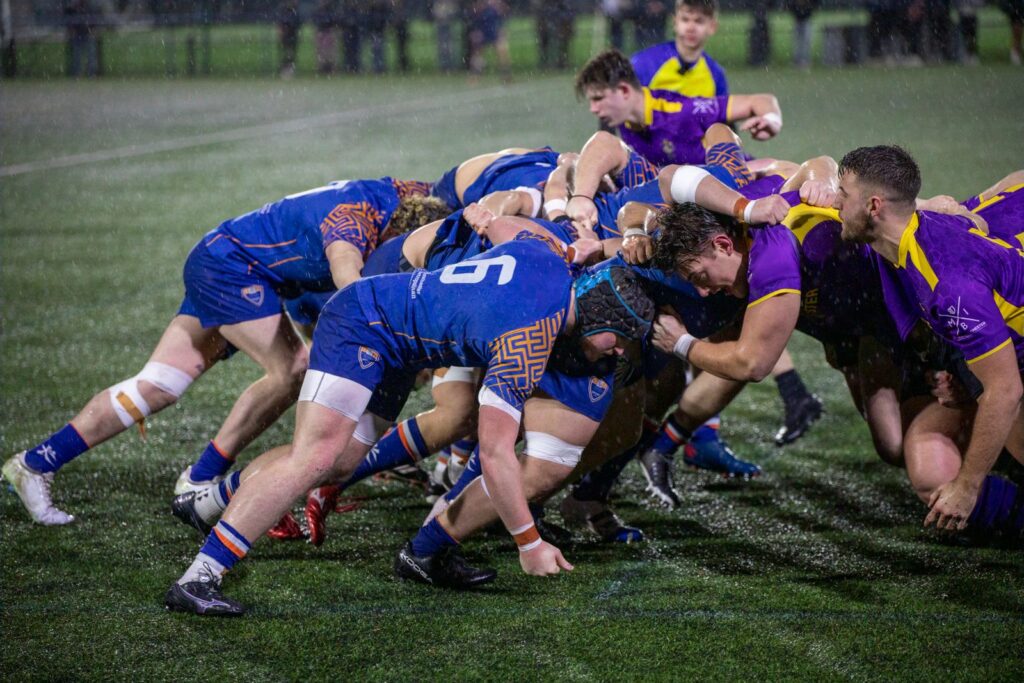 Two rugby packs in blue and in purple bound together in a scrum on a wet evening pitch
