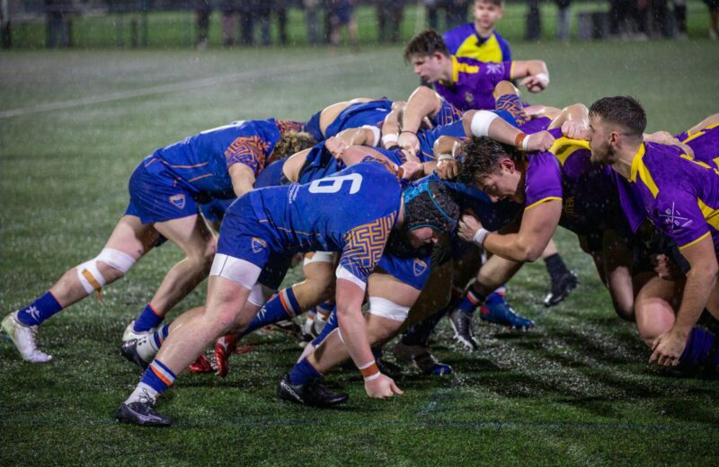 Two rugby packs in blue and in purple bound together in a scrum on a wet evening pitch