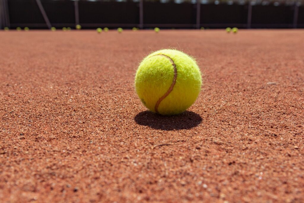 A yellow tennis ball on a red clay court, the court lines blurred behind it