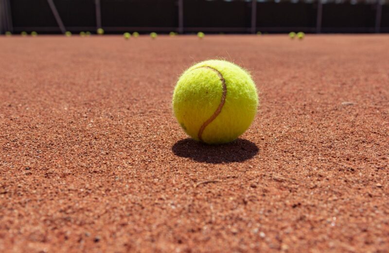 A yellow tennis ball on a red clay court, the court lines blurred behind it