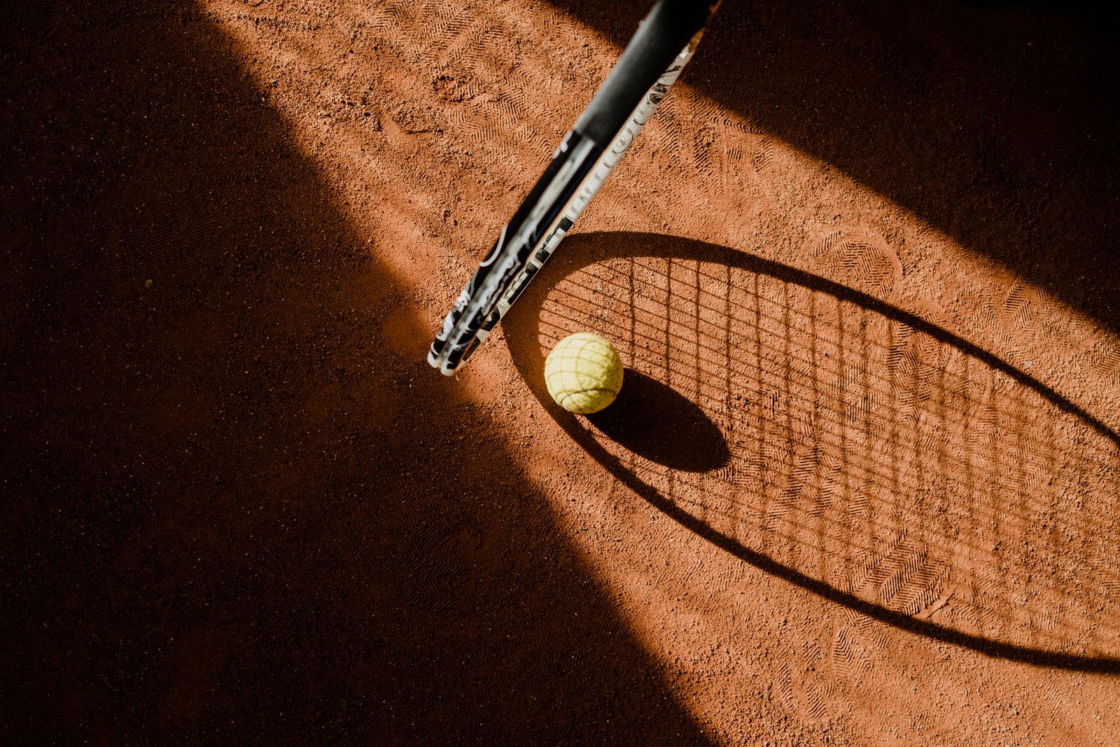 The shadow of a tennis racket and a ball on a sunlit clay court