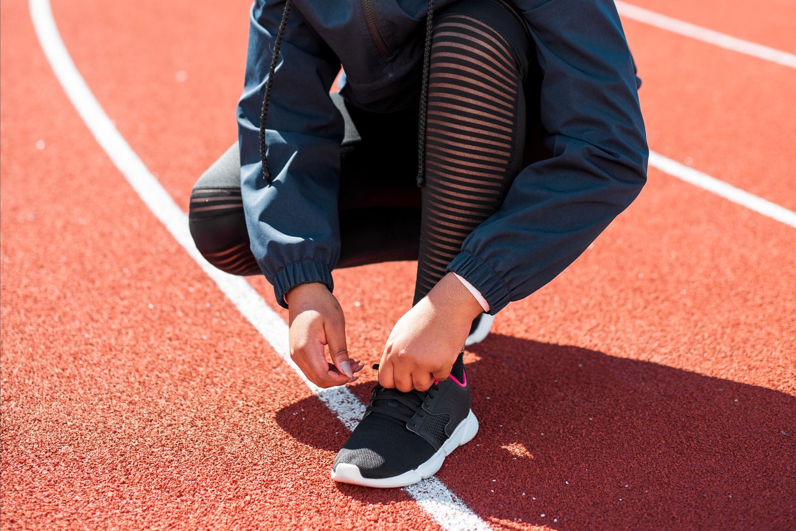 A runner in a navy jacket and black tights crouching on a red running track to tie a black running shoe