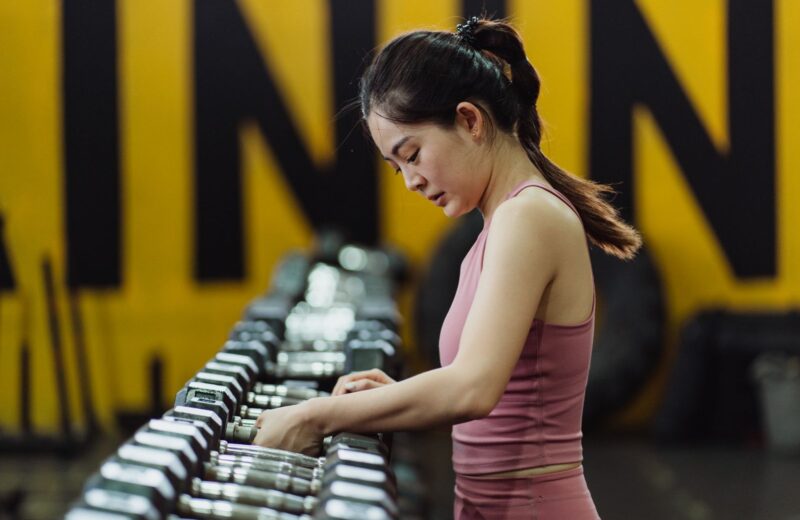 A woman in a pink training top choosing a dumbbell from a long rack in a gym