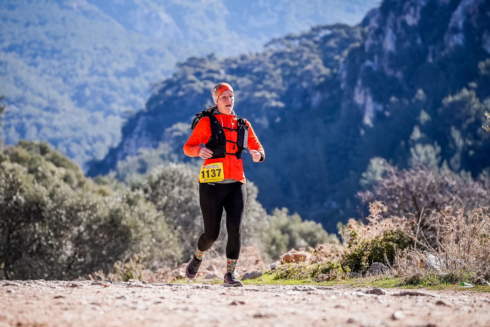 A trail runner in an orange jacket, running vest and tights on a stony path, with wooded mountains behind