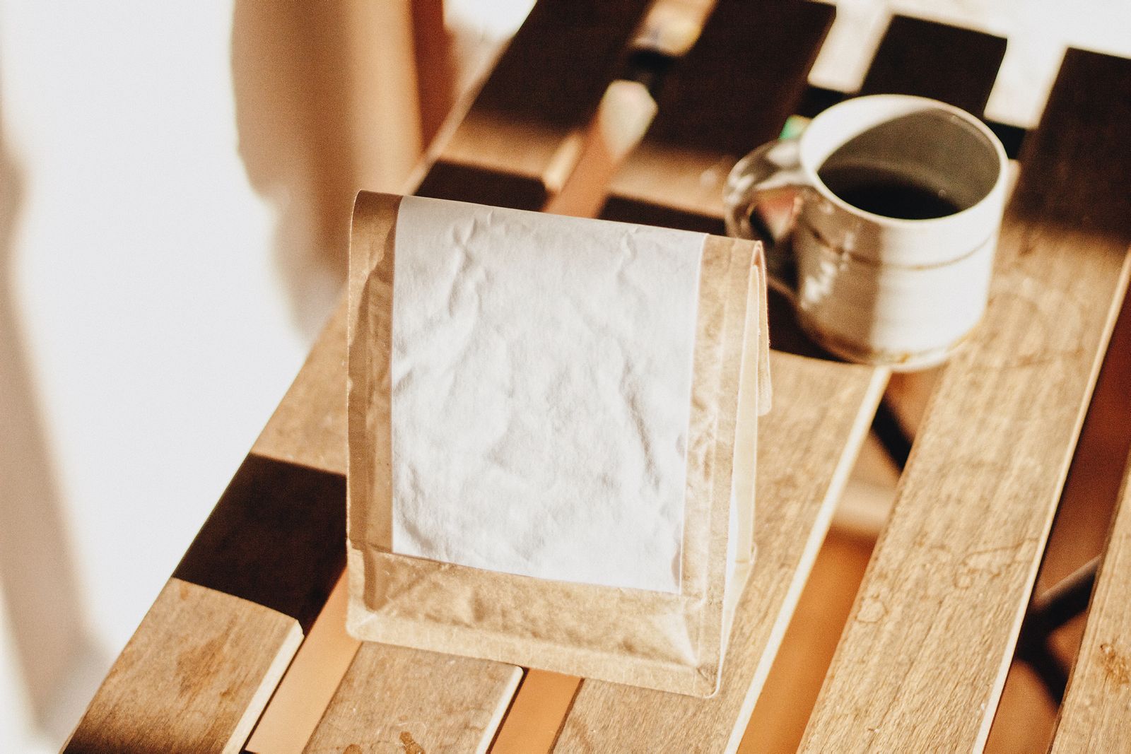 A plain paper coffee bag and a mug on a sunlit wooden table