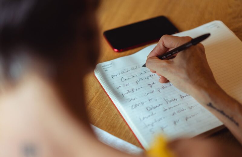 A person writing a handwritten list in a notebook at a wooden table