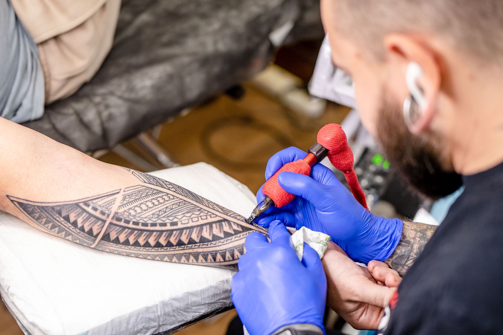 An artist in blue gloves tattooing a black patterned piece along a client's forearm