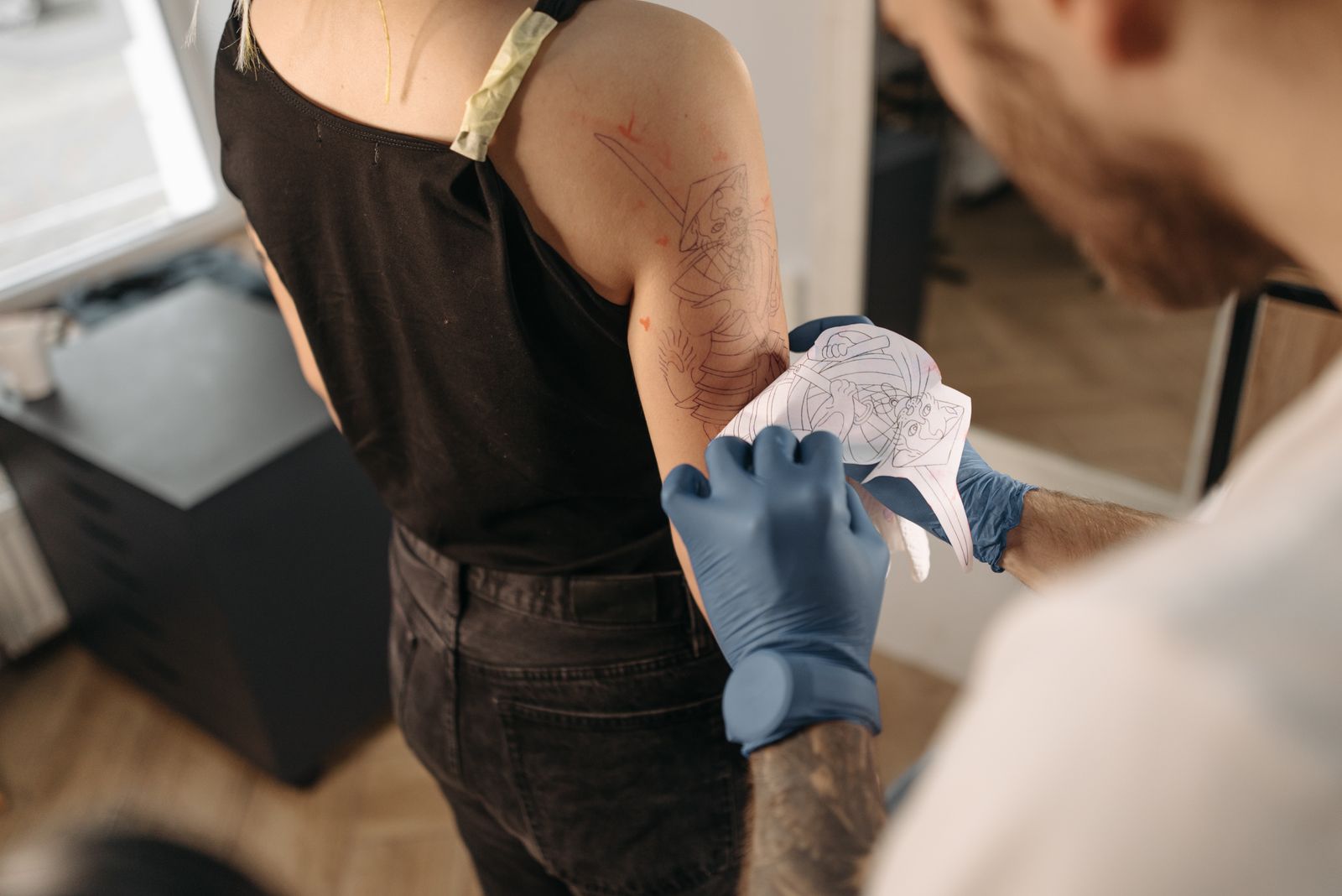 An artist in blue gloves pressing a paper stencil onto a client's upper arm