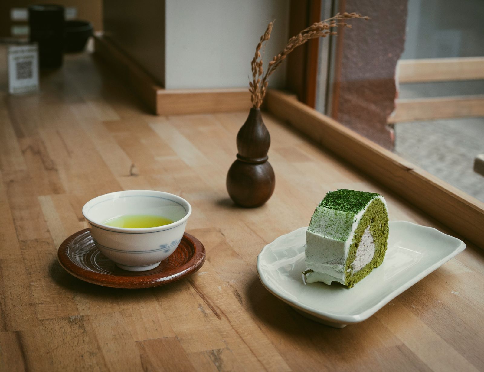 A bowl of green tea and a slice of matcha roll cake on a wooden table by a window