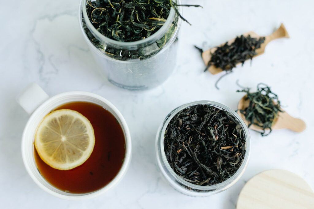 Jars of dark loose-leaf tea beside a cup of tea with lemon on white marble