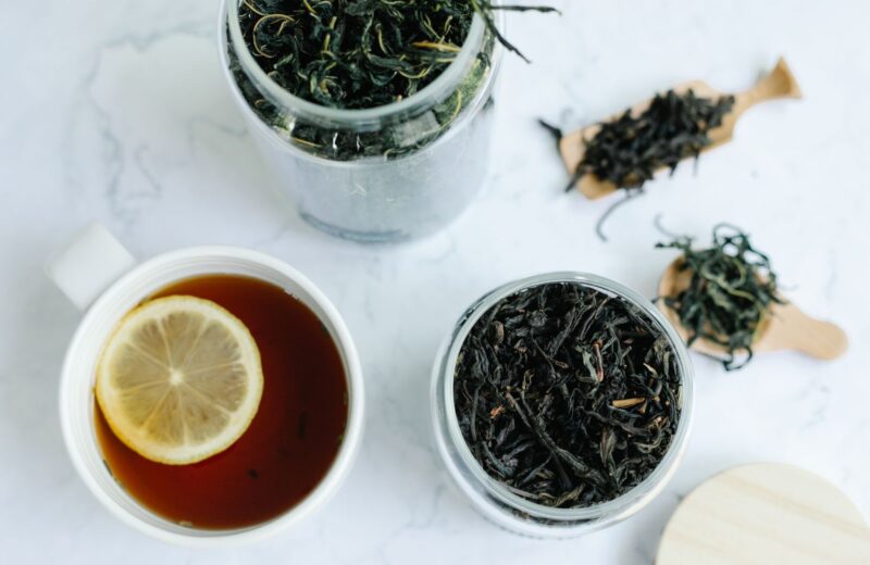 Jars of dark loose-leaf tea beside a cup of tea with lemon on white marble