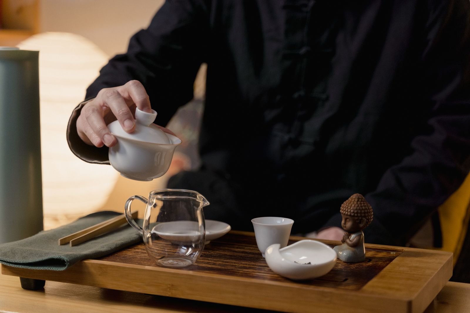 Hands pouring tea from a small porcelain pot into a glass pitcher on a wooden tea tray