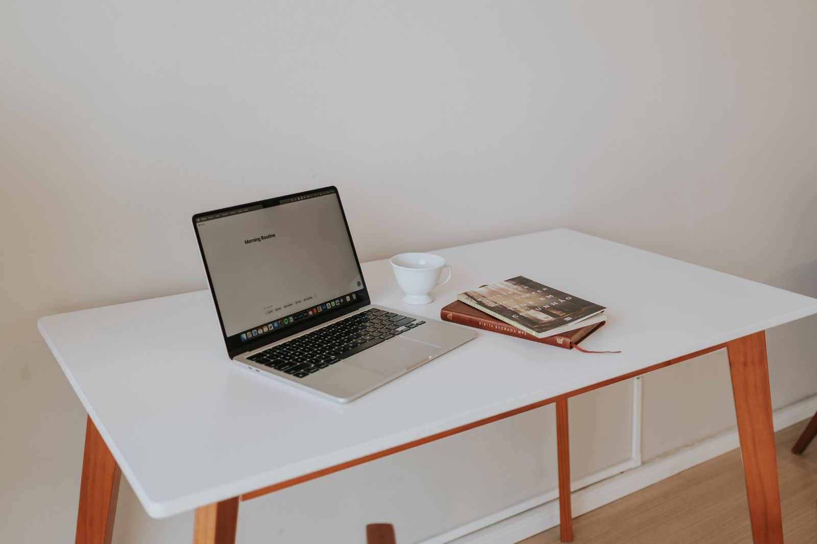 An open laptop on a small white desk with a cup and two books
