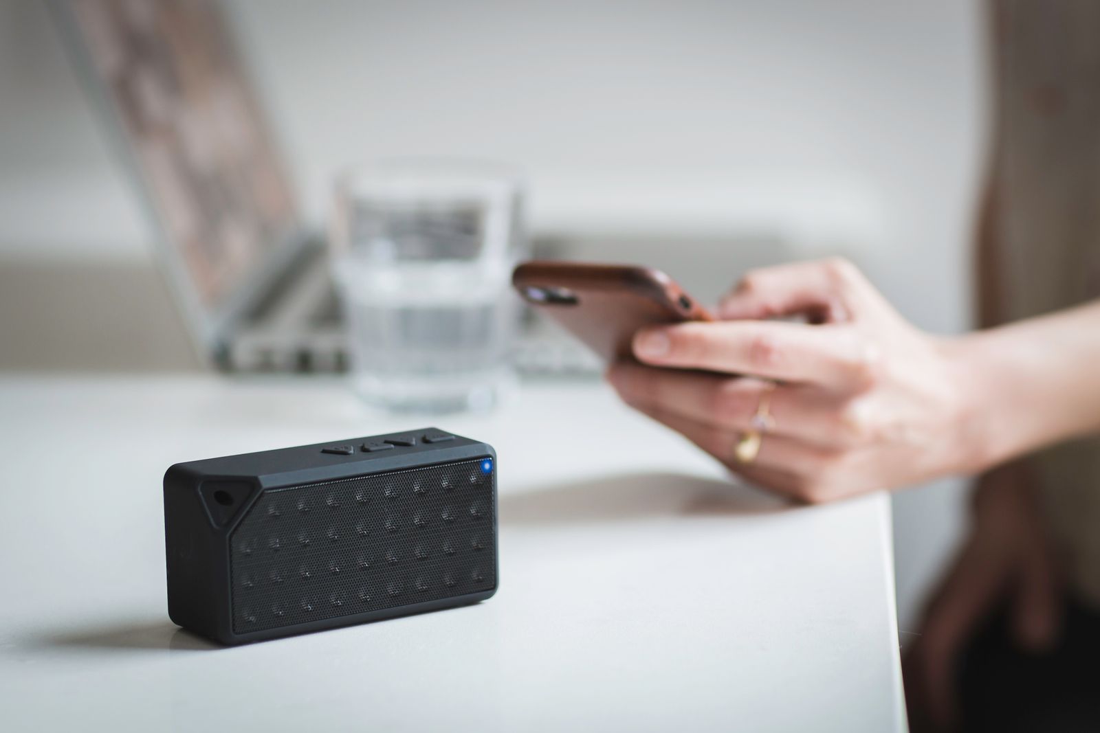 A small black portable speaker on a white table while a hand scrolls a phone beside it