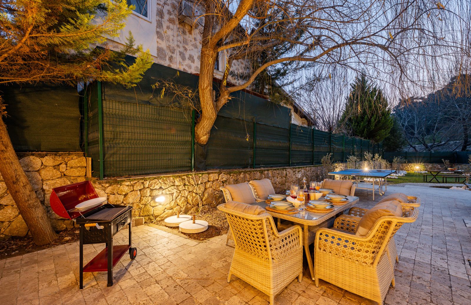An outdoor dining table with wicker chairs and a grill on a lit stone terrace at dusk