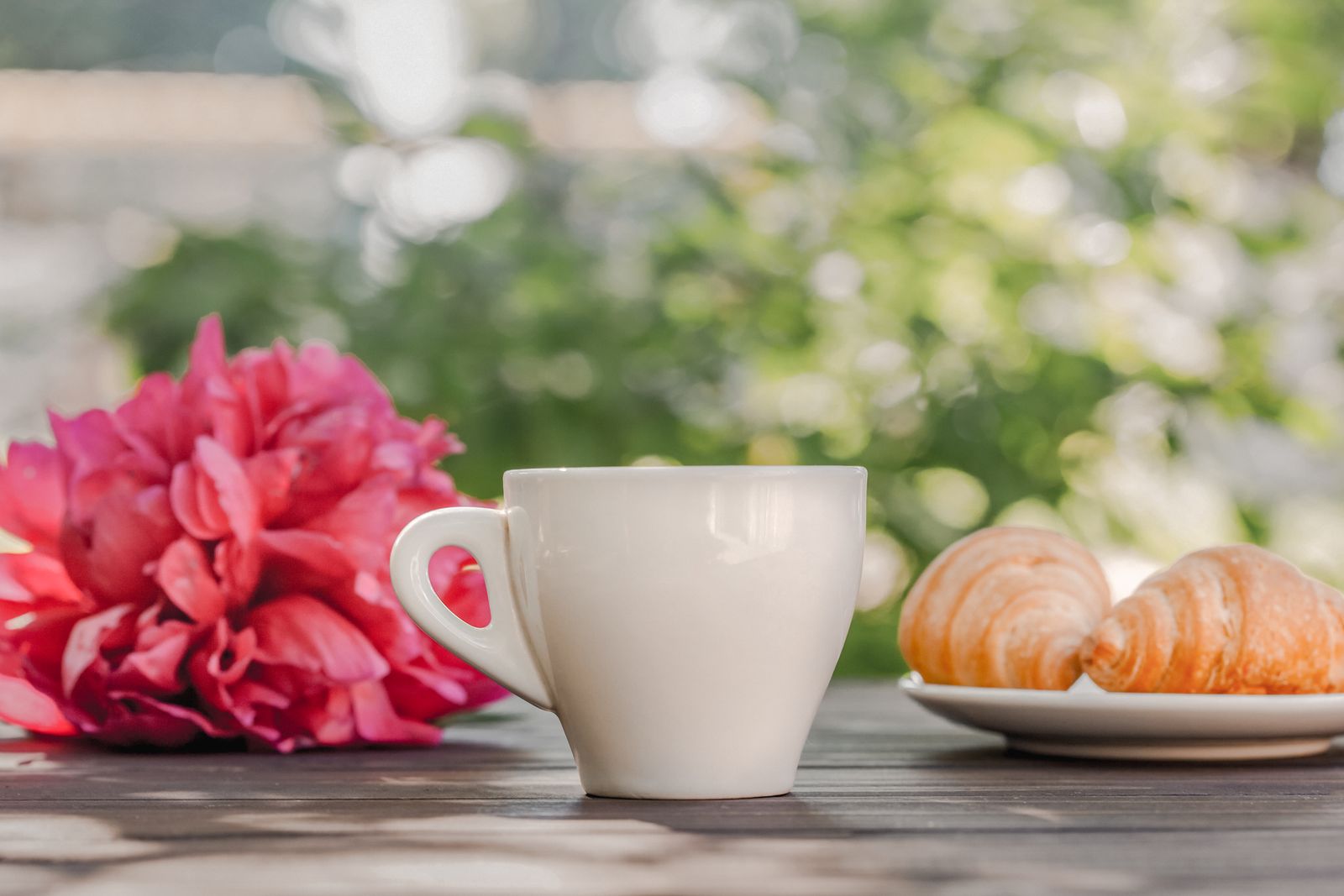 Coffee and croissants on a wooden terrace table in morning sun, with pink peonies