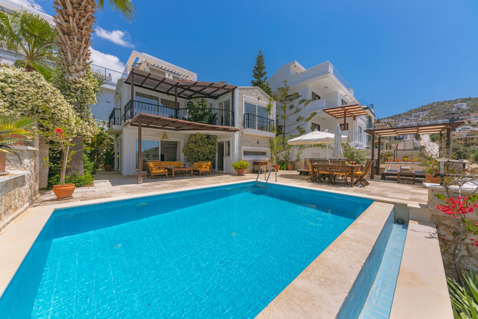 A long blue swimming pool beside a white villa with a terrace, palm trees and loungers under a clear sky