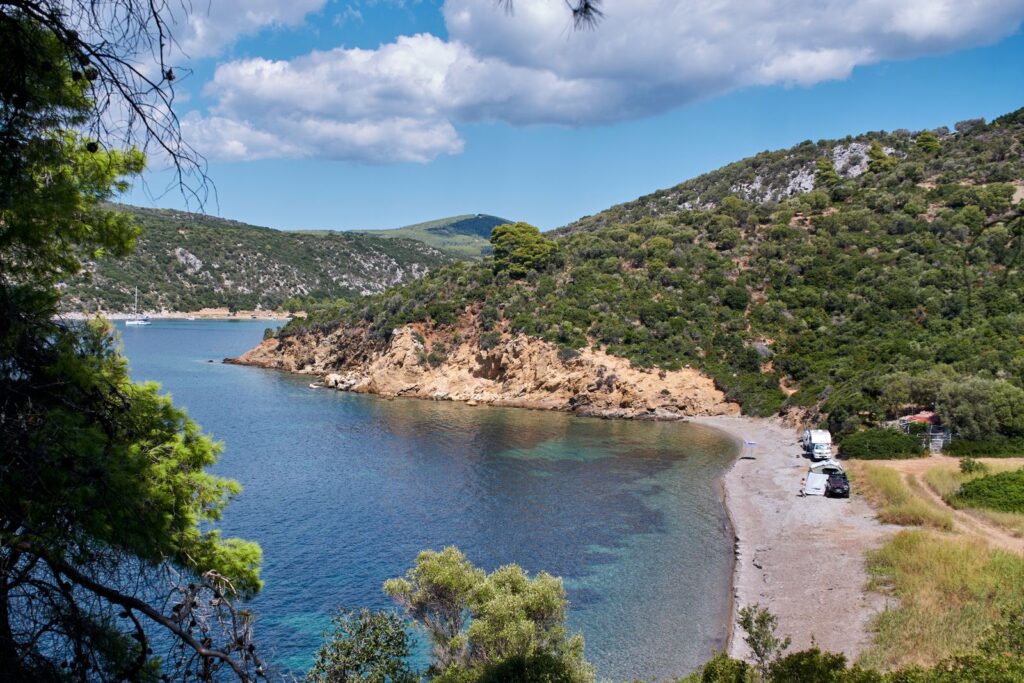 A quiet bay with green hills and a pebble beach seen through the trees