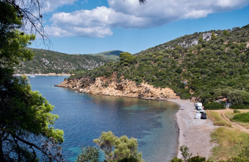 A quiet bay with green hills and a pebble beach seen through the trees